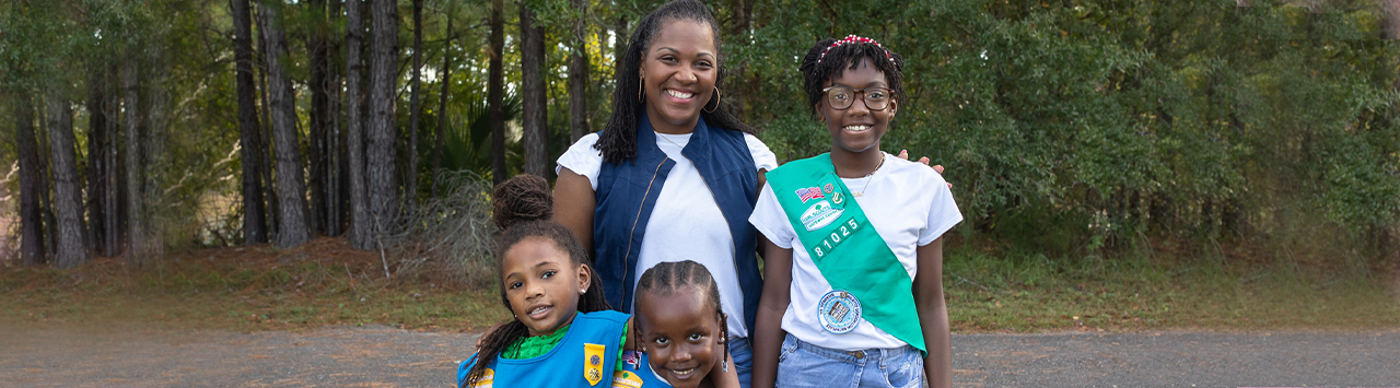  adult volunteer wearing vest with a girl scout junior (right, in baseball cap) and daisy (left) outside at a park smiling and looking at one another 